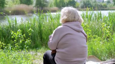 Nehir manzarasına bakan, kütüğün üzerinde oturan yaşlı bir kadının arka görüntüsü. Emekliler mavi gökyüzünün, yeşil ağaçların, sazlıkların, gün ışığındaki nehrin manzarasının tadını çıkarırlar. Dinlenme, rahatlama, sıcak hava. Gün batımı.