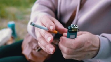 Elinde sigara tutan kadın ve doğada güneşin altında oturan bir çakmak. Dışarıda sigara içen bir kadın, park eder. Sigara içiyorum. Kötü alışkanlık. Bağımlılık. Sağlıksız yaşam tarzları. Stres yüzünden. Dinleniyor.