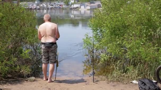 漁師はリールで回転ロッドを使用して水からキャッチ魚を引き出します バックビュー 裸の胴と2本のロッドを持つ男性は 日没時に日光の光の中で池で釣りをしています スポーツ レクリエーション — ストック動画