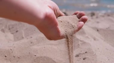 Female Hand Pours Sand Through Fingers on the Beach in the Rays of Sunlight. Fine grains of sand slowly fall on the hot golden sand in the sun glare at sunset. Rest on the sea coast, a seaside resort.