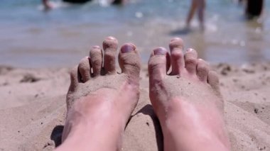 Sand Lies on a Sandy Beach of the Ocean 'da bronzlaşmış Kızıl Çıplak Kadın Ayakları. Kız güneşin altında ayak parmaklarını hareket ettiriyor. Bacakları güneşin parlaklığında güneşleniyor. Yüzen turistler. Dinlen, dinlen..