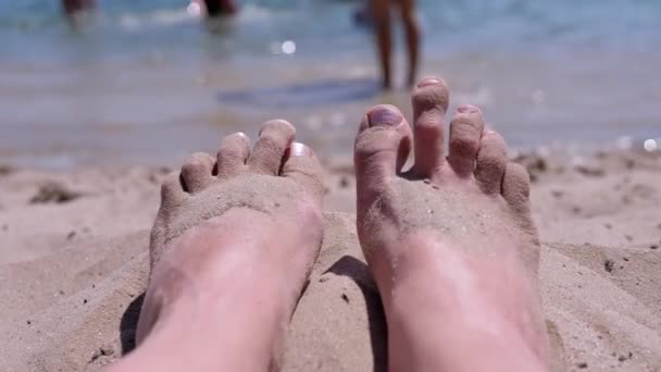 在海洋海岸的沙滩上 晒黑的赤裸女脚躺在沙滩上 女孩在烈日下 脚趾在阳光下移动 腿在阳光下晒日光浴 飘浮的游客 — 图库视频影像