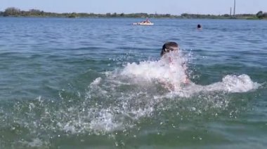 Happy Child Swimming, Splashing on the Beach in Sunlight on Sea. Boy playing in the water, creates waves, splashing with hands. Family rest, recreation, summer vacation. Sea resort. Weekend. Summer.