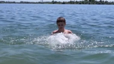 Üzgün Çocuk Yüzme, Denizde Güneş Sahilinde Su Sıçratma. Çocuk suda oynuyor, dalgalar yaratıyor, ellerle su sıçratıyor. Aile dinlenmesi, dinlenme, yaz tatili. Deniz tatil köyü. Hafta sonu. Yaz zamanı..