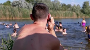 Plajda oturan erkek Bulanık Nehir Arkaplanı 'nda Smartphone' da konuşuyor. Genç bir adam sazlıkların yakınındaki gölde yüzen çocuklara bakıyor, doğanın tadını çıkarıyor, güneş ışığında deniz manzarası seyrediyor. Hafta Sonu.