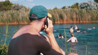 Plajda oturan erkek Bulanık Nehir Arkaplanı 'nda Smartphone' da konuşuyor. Şapkalı genç bir adam sazlıkların yakınındaki gölde yüzen çocuklara bakıyor, doğanın tadını çıkarıyor, güneş ışığında deniz manzarası seyrediyor..
