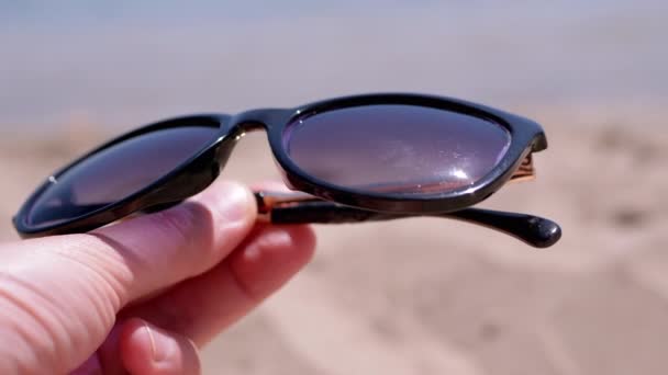 女性手拿着深色太阳镜 靠着海景的背垂 女孩在热带海岸的沙滩上享受着放松的生活 太阳光反射在眼镜上 夏天的时间 — 图库视频影像