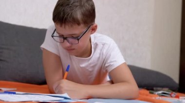 Not Defterinde Kalemle Yazan Çocuk Odadaki Turuncu Yatak Örtüsü Üzerine Yatan Çocuk. Gözlüklü zeki bir genç rahat bir pozisyonda yerde ödev yapıyor. Eğitim, öğrenme kavramı.
