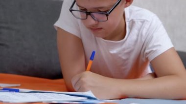 Child Writing with a Pen in a Notebook, Lying on an Orange Bedspread in the Room. A serious smart teenager in glasses is doing homework on floor in a comfortable position. Education, learning concept.
