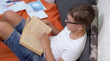 Gözlüklü Zeki Çocuk Odada Yerde Oturan İlginç Bir Kitap Okuyor. Yan görüş. Ciddi bir çocuk sayfaları eliyle çevirir. Ödev yapan, ders çalışan özenli bir öğrenci. Öğrenme.