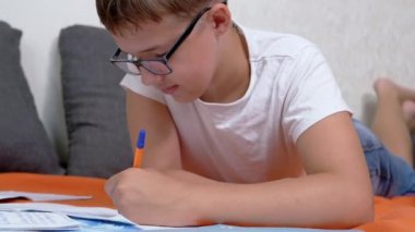 Not Defterinde Kalemle Yazan Çocuk Odadaki Turuncu Yatak Örtüsü Üzerine Yatan Çocuk. Gözlüklü zeki bir genç rahat bir pozisyonda yerde ödev yapıyor. Eğitim, öğrenme kavramı.