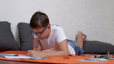 Not Defterinde Kalemle Yazan Çocuk Odadaki Turuncu Yatak Örtüsü Üzerine Yatan Çocuk. Gözlüklü zeki bir genç rahat bir pozisyonda yerde ödev yapıyor. Eğitim, öğrenme kavramı.