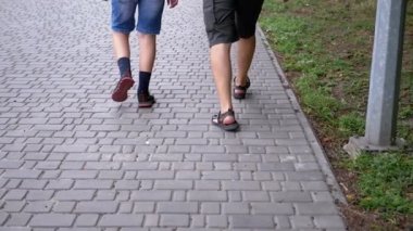 UKRAINE, KAMENSKOE, 2022: Legs of Father and Son Walking on Sidewalk Outdoors in Park near Green Grass. Close-up. Slow-motion. Two people, a male, and a teenager are walking together on the street.