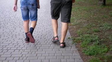 UKRAINE, KAMENSKOE, 2022: Legs of Father and Son Walking on Sidewalk Outdoors in Park near Green Grass. Close-up. Slow-motion. Two people, a male, and a teenager are walking together on the street.