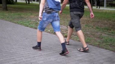UKRAINE, KAMENSKOE, 2022: Legs of Father and Son Walking on Sidewalk Outdoors in Park near Green Grass. Close-up. Slow-motion. Two people, a male, and a teenager are walking together on the street.