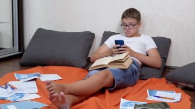 Gözlüklü, elinde akıllı telefon olan ve internette kitap okuyan bir genç. Akıllı çocuk, dağınık kitaplar ve defterler arasında turuncu bir yatak örtüsüne uzanmış bacaklarını uzattı. Eğlence. Eğitim.