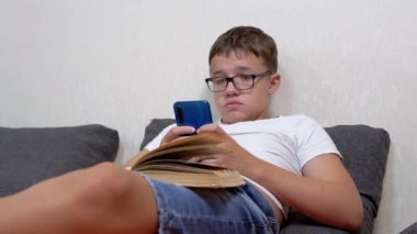 Teen in Glasses with a Smartphone in Hands and Open Book Studying Online at Home. Serious smart teenager writes a message, looks at a mobile app. Lessons. Entertainment. Education. Games. Resting.
