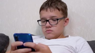 Tired Teenager in Glasses Plays on a Mobile Phone While Lying on Pillow in Room. A serious smart boy reads, writes a message, looks at the app on the smartphone screen. Child studying online at home.