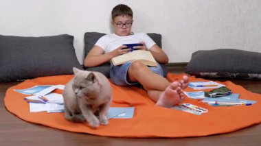 Smiling Teenager in Glasses Reading Messages on a Mobile App on a Smartphone. Tired schoolboy plays on a mobile phone while lying on a pillow on orange rug together with a cat, among scattered books.