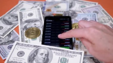 Broker is Viewing the Price Change of a Cryptocurrency on a Smartphone Screen. Rising, falling price. Dollar bills, bitcoin coins are scattered on a table. BTC, LTC, USD in app online. Stock exchange.