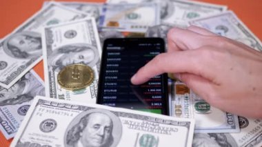 Broker is Viewing the Price Change of a Cryptocurrency on a Smartphone Screen. Rising, falling price. Dollar bills, bitcoin coins are scattered on a table. BTC, LTC, USD in app online. Stock exchange.