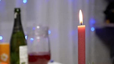 Burning Red Christmas Candle Burns with a Bright Flame on the New Years Table. Christmas decorated table on eve of holiday on background bulbs garlands. Cozy family dinner in a festive atmosphere.