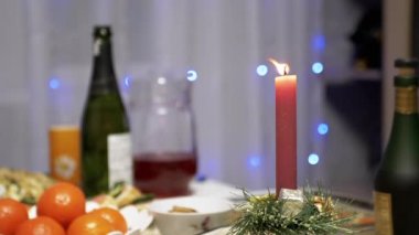 Burning Red Christmas Candle Burns with a Bright Flame on the New Years Table. Christmas decorated table on eve of holiday on background bulbs garlands. Cozy family dinner in a festive atmosphere.
