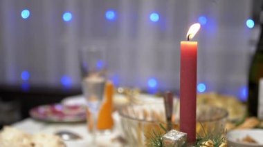 Burning Red Christmas Candle Burns with a Bright Flame on the New Years Table. Christmas decorated table on eve of holiday on background bulbs garlands. Cozy family dinner in a festive atmosphere.