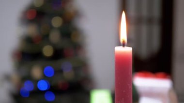 Burning Red Candle with a Bright Flame on a Blurred Christmas Tree Background. Decorated Christmas tree in the background with blurry spots of colorful garland. A new year at home. Winter holiday. 4K.