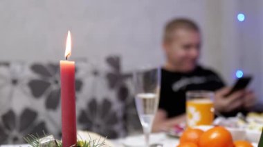 Burning Red Christmas Candle Burns with a Bright Flame on the New Years Table. Male is resting with a smartphone on a blurred background of a twinkling garland. A new year at home. Winter holiday.