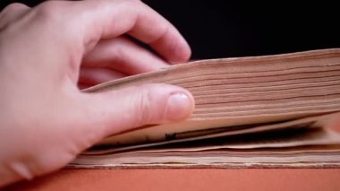Female Fingers Leafing Through Pages of Old Shabby Book on a Black Background. Reading. Rough papyrus paper on a thick antique book from yesteryear. Orange color. Education. Knowledge. Mystic.