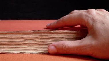 Female Fingers Leafing Through Pages of Old Shabby Book on a Black Background. Reading. Rough papyrus paper on a thick antique book from yesteryear. Orange color. Education. Knowledge. Mystic.