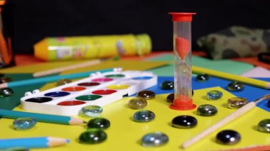Hourglass on the Background of Scattered Multi Colored Cardboard, School Supplies. Time lapse. Time concept. Flowing sand. End of lesson. Creativity, homework. Yellow background. Childhood. Education.