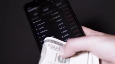 Businessman is Viewing Quotes Data, Price of a Cryptocurrency on a Smartphone. Broker holds a smartphone in his hands, 100 dollar bills, analyzes the price index on the stock market. Black background.