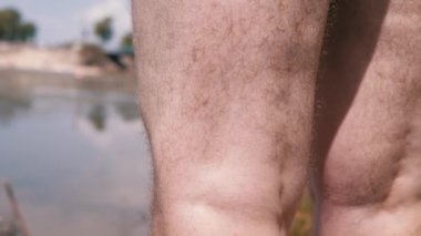 Male Hairy Curly Legs Standing on Sand near Water in Rays of Sunlight on Nature. An unrecognizable barefoot man is resting on bank of the river, enjoying the scenery at sunset. Body part. Back view.