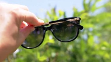 Female Hand Stretches Dark Sunglasses Against the Backdrop of Nature. Girl enjoying, relaxing in leafy forest. Sun glare is reflected in lenses. Blurred background of trees, sky. Summertime. Weekend.