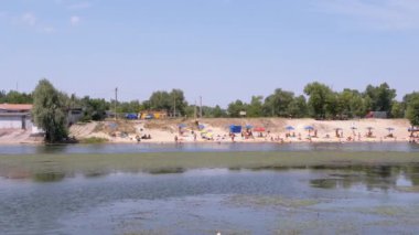 Panoramic View of City Beach with Umbrellas, People Resting on the River Bank. Coastline, blooming green river, beach area. Blue sky, white sand, bathing tourists. Nature, summer landscape. Weekend.
