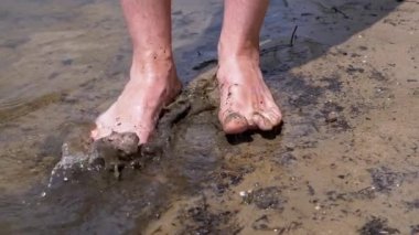 Male Feet are Splashing in Mud, Dirty Water, Standing on the River Bank on Sun. Bare male feet step into a small dirty puddle, splashing drops of water around. Fun, play. Rest, weekend on nature.