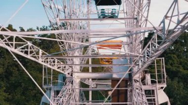 Mechanism of a Rotating Old Metal Ferris Wheel, View from Inside the Cabin. Rising wheel above city, urban landscape view, blue sky, green trees, rooftops. Scenery at sunset. Amusement park. Leisure.