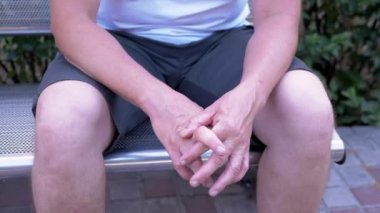 Male Rubbing Hands Sitting on a Bench in the Urban Park. A moment of waiting, excitement, anxiety, and emotional stress, trouble, nerves, and dread. Massage of hands, joints, fingers, skin. Outdoors.