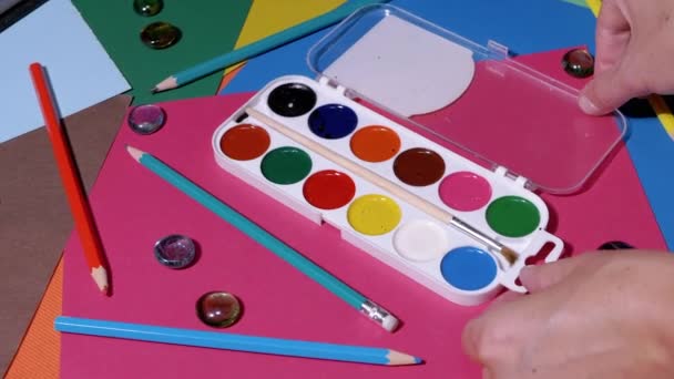 女性の手図面のテーブルから水彩のパレットを削除します 色ボール紙 紙の散乱色鉛筆の背景に多色の塗料のセット 職場清掃 — ストック動画