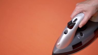 Ütü masasında Buhar Ütülü Parlak Turuncu Kumaşı Ütüleyen Kadın El. Yakın plan. Rutin ev işleri. Ev sahibesinin görevleri. Turuncu arka plan. Tekstil. Modern buhar makinesi, buhar ütüsü. Yavaş çekim.