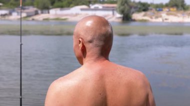 Angler Standing on Riverbank, Catching Fish, Looking into the Distance on Sunset. Back view of a bald man enjoying a fishing trip. Leisure, hobby, lifestyle. Nature, summer landscape. Weekend.