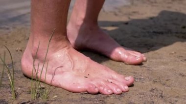 Male Tanned Legs Standing on Wet Sand near Water in Rays of Sunlight in Nature. Unrecognizable barefoot man resting on bank of the river enjoying the scenery at sunset. Nature in the woods. Weekend.