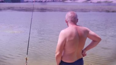 Angler Standing on Riverbank, Catching Fish, Looking into the Distance on Sunset. Back view of a bald man enjoying a fishing trip. Leisure, hobby, lifestyle. Nature, summer landscape. Weekend.