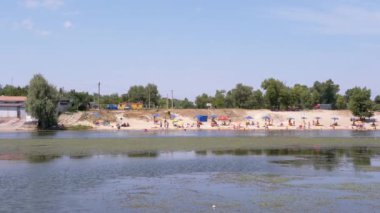 Panoramic View of City Beach with Umbrellas, People Resting on the River Bank. Coastline, blooming green river, beach area. Blue sky, white sand, bathing tourists. Nature, summer landscape. Weekend.