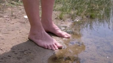 Male Tanned Legs Standing on Wet Sand near Water in Rays of Sunlight in Nature. Unrecognizable barefoot man resting on bank of the river enjoying the scenery at sunset. Nature in the woods. Weekend.
