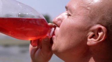 Bald Wrinkly Man Greedily Drinks Juice, Drink, Colored Water Against the Sky. Man drinks a red drink from a plastic bottle in the rays, glare of sunlight. Thirst in nature, on the beach. Summertime.