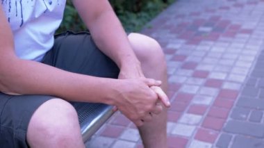 Male Rubbing Hands Sitting on a Bench in the Urban Park. A moment of waiting, excitement, anxiety, and emotional stress, trouble, nerves, and dread. Massage of hands, joints, fingers, skin. Outdoors.