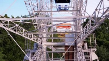 Mechanism of a Rotating Old Metal Ferris Wheel, View from Inside the Cabin. Rising wheel above city, urban landscape view, blue sky, green trees, rooftops. Scenery at sunset. Amusement park. Leisure.